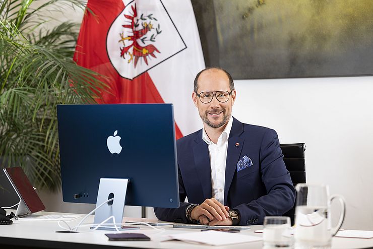 Mann sitzt vor Computer am Tisch, im Hintergrund eine Tirol-Fahne
