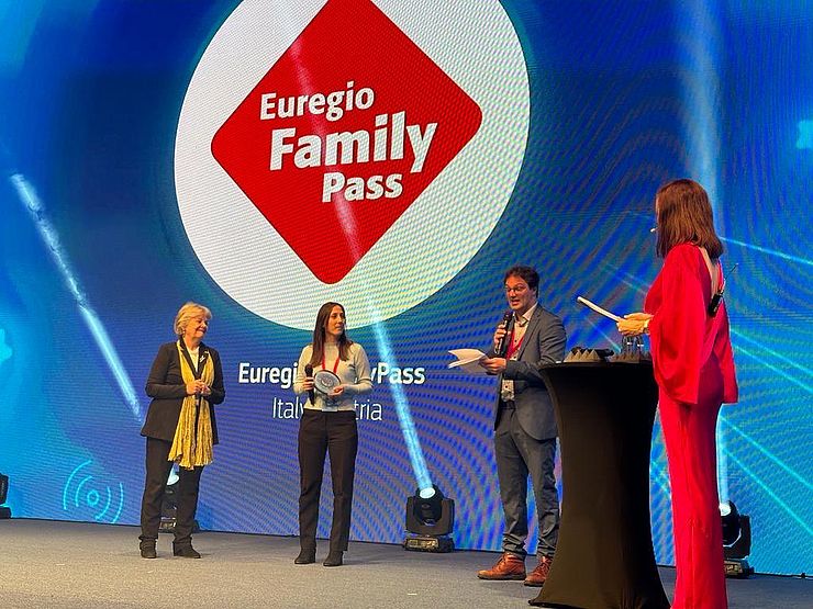 Vor Bildschirm mit Logo EuregioFamilyPass stehen Moderator und die beiden Stellvertreterinnen der Euregio