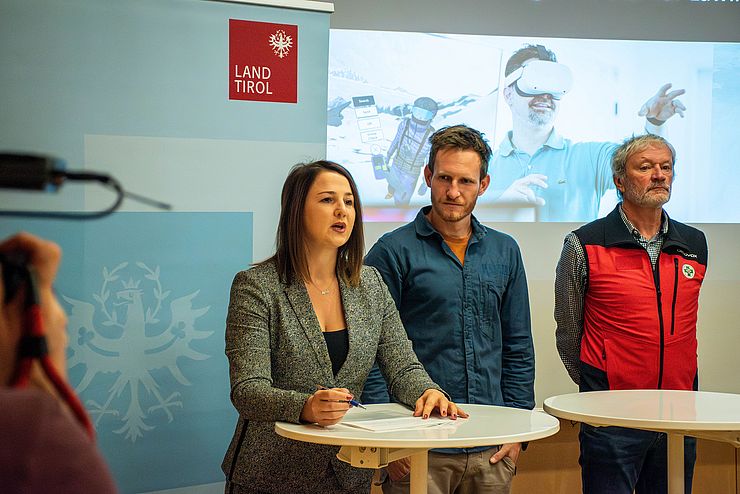 LRin Mair, Lanzanasto und Spiegl stehen vor Stehtischen; LRin Mair spricht; im Hintergrund ein Roll-Up mit Land Tirol Logo und eine Leinwand auf der ein Bild des Simulators zu sehen ist