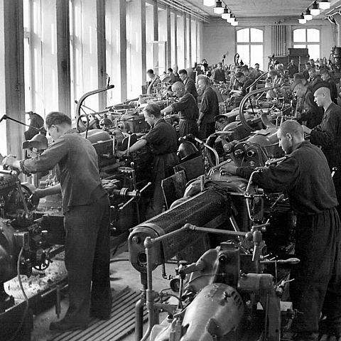 In einheitlicher Arbeitskleider stehen jeweils Männer vor einer Drehbank in einer Fabrikshalle.