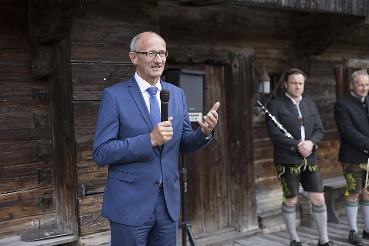 MAttle mit blauen Anzug und Krawatte ein Mikrofon in der Hand; m Hintergrund eine Wand einer Holzhütte; am rechten Bildrand zwei Musiker in Lederhose mit Instrumenten in der Hand