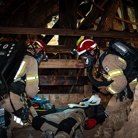 Dunkler Stollen: zwei Mitglieder der GRubenwehr mit Helm und Atemschutzmaske knieen über einer am Boden liegenden Person