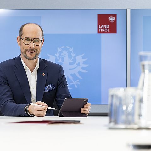Mario Gerber sitzt am Tisch und hält Tablet in der Hand, im Hintergrund Bildschirm mit Land Tirol Logo