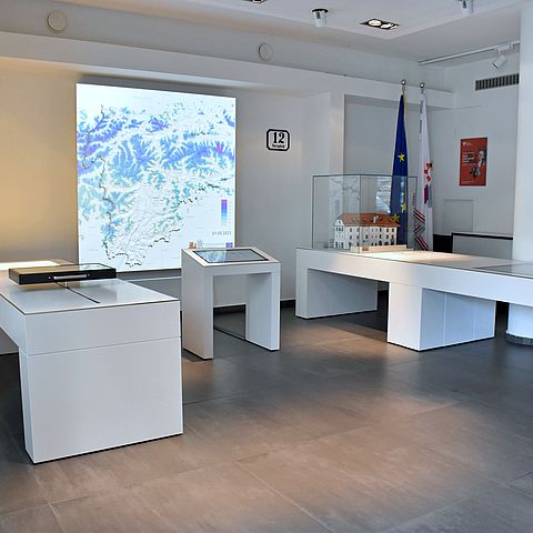 Auf dem Bild ist die Ausstellung zu sehen. Man kann drei weiße Tische mit Bildschirmen und im Hintergrund eine Karte der Europaregion Tirol-Südtirol-Trentino erkennen. 