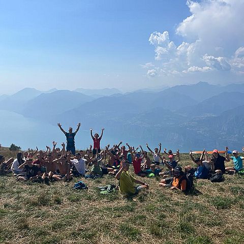 Gruppe Jugendlicher streckt Hände nach oben, sitzt auf Wiese - dahinter Bergkulisse und See