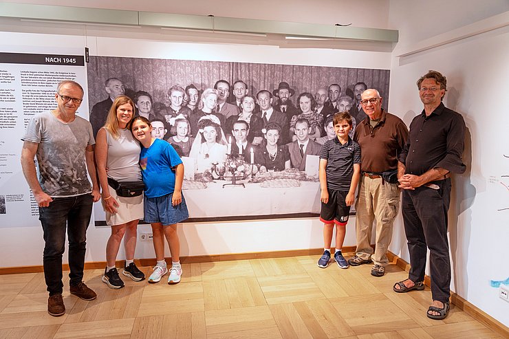 © Land Tirol/Saxl Sechs Personen stehen vor einem großen Familienportrait an einer Wand.