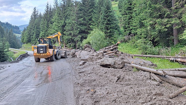 Bagger befreit Straße von Erde/Schlamm mit Steinen und Baumstämmen.