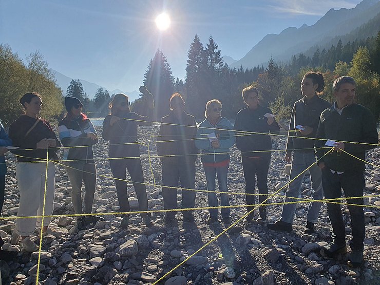 Mehrere Menschen stehen auf einem steinigen Flussbett und halten gelbe Schnüre in den Händen, die zwischen ihnen gespannt sind und ein Netz bilden. Einige halten ebenso kleine Karten in den Händen. Im Hintergrund sind Bäume, ein Fluss, Berge und die tief stehende Sonne zu sehen.