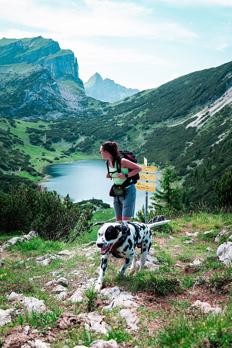 Eine Frau mit Hund auf einem Wanderweg im Hintergrund ein Bergsee und gelbe Hinweisschilder