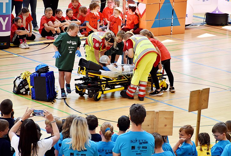 Ein Sanitäter und eine Sanitäterin heben gemeinsam mit Kindern eine verletzte Person auf eine Trage.