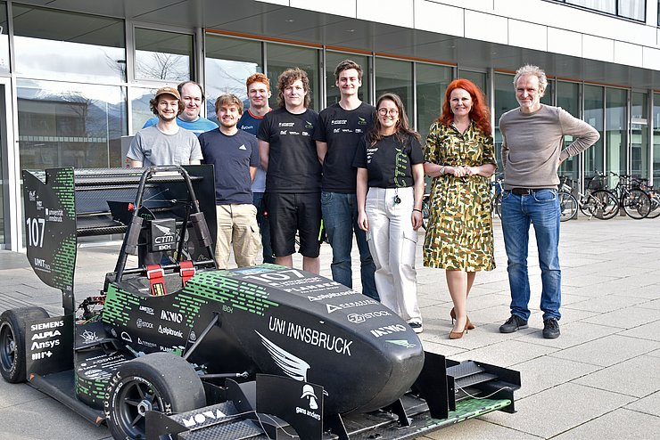 Gruppenfoto von mehreren Personen die vor einem Gebäude stehen, im Vordergrund ist ein Rennauto zu sehen