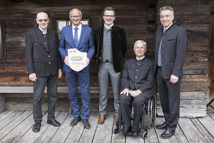 Foto mit LH MattleMattle mit einem Schild aus Holz in der Hand (Wappen Tirol/Bayern, Zeichnung der neuen Innfähre, Aufschrift Europatag 2023), Schneider, Gruber, der im Rollstuhl sitzt und Lederer; im Hintergrund Wand eines Holzhauses und eine Holzbank