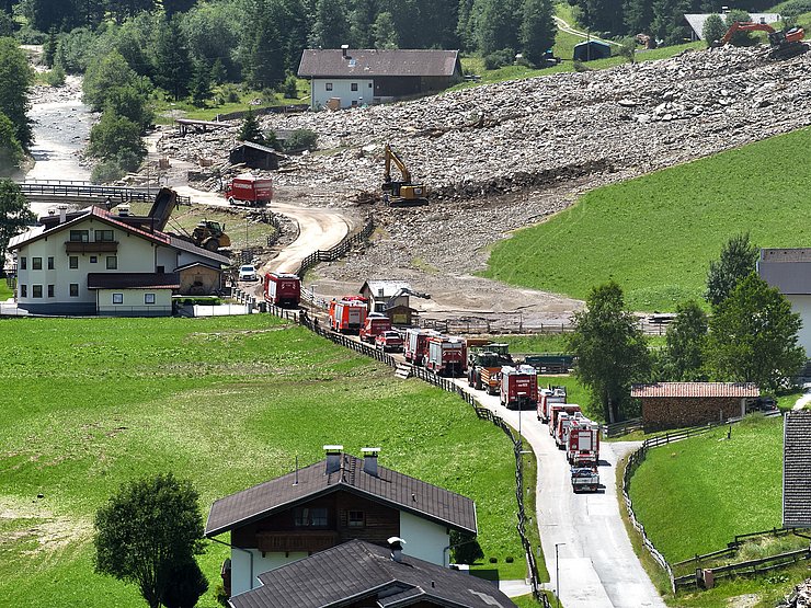 © Land Tirol/Drohnen-Einsatz-Gruppe Blick auf Talboden. Im hinteren Bereich sind ein Haus und Ablagerungen eines Murgangs zu sehen, die mit zwei Baggern bearbeitet werden. Im Vordergrund stehen Einsatzfahrzeuge der Feuerwehr auf einer Straße. Links und rechts davon sind Häuser und Wiese zu sehen.