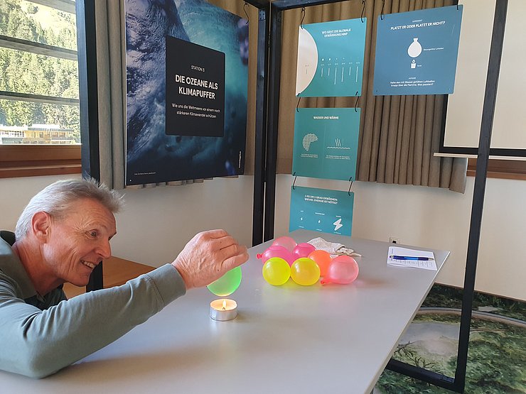 Ein älterer Mann sitzt an einem Tisch und hält einen kleinen Luftballon über eine brennende Kerze. Auf dem Tisch liegen weitere kleine Luftballons. Im Hintergrund hängen Informationstafeln mit der Aufschrift „Die Ozeane als Klimapuffer“ und weiteren Informationen zum Thema.