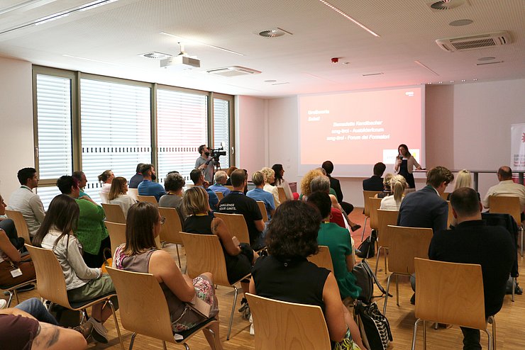 Saal mit Personen auf Stühlen, von hinten - vorne die Bühne und Rednerin