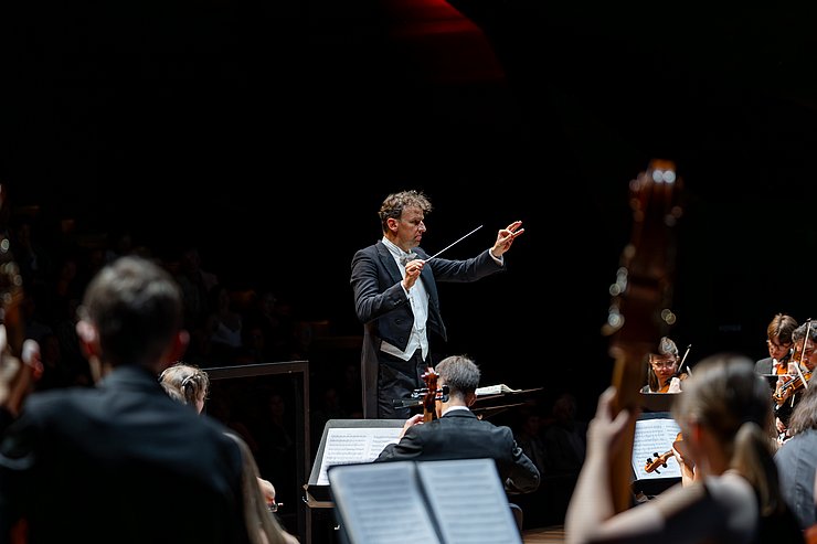 Ein Mann in Frack dirigiert ein Orchester. 