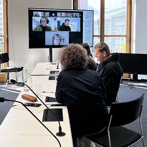 VertreterInnen sitzen im Medienraum, Bildschirm zeigt andere Online-TeilnehmerInnen