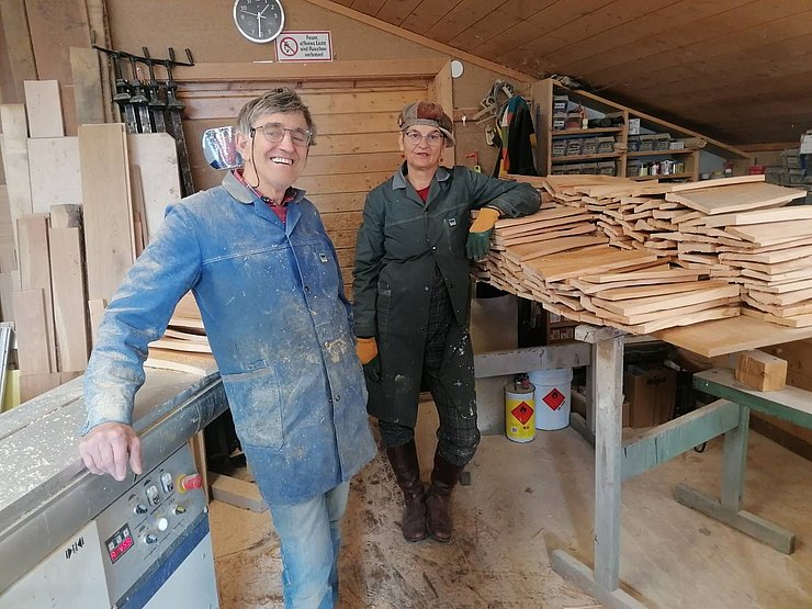 Zwei Personen stehen in einer Holzwerkstatt. Beide tragen Arbeitskleidung. Auf einer Maschine und einem Arbeitstisch liegen Holzschindeln. Im Hintergrund Werkzeug, Holzleisten und Regale mit Materialien.