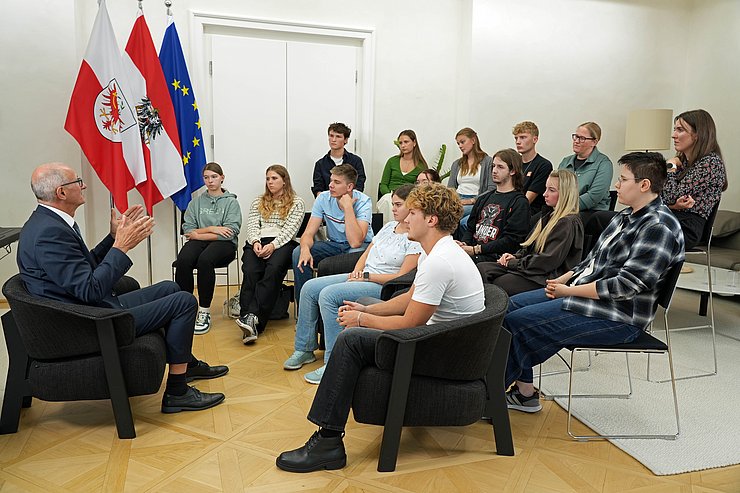 © Land Tirol/ Saxl Landeshauptmann Anton Mattle sitzt in einem Sessel und unterhält sich mit einer Schulklasse. Die Jugendlichen sitzen im Halbkreis um ihn und hören ihm aufmerksam zu. Im Hintergrund stehen die Flaggen von Tirol, Österreich und der EU.