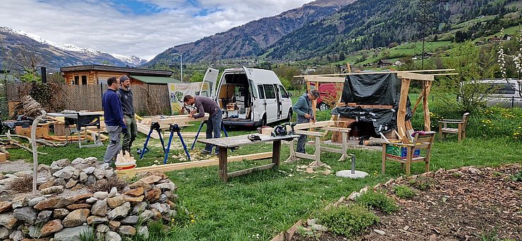 Mehrere Personen arbeiten auf einer Wiese des Gemeinschaftsgartens mit Holz für das Schindeldach. In der Mitte steht ein geöffneter weißer Kleintransporter mit Werkzeug und Material im Laderaum.