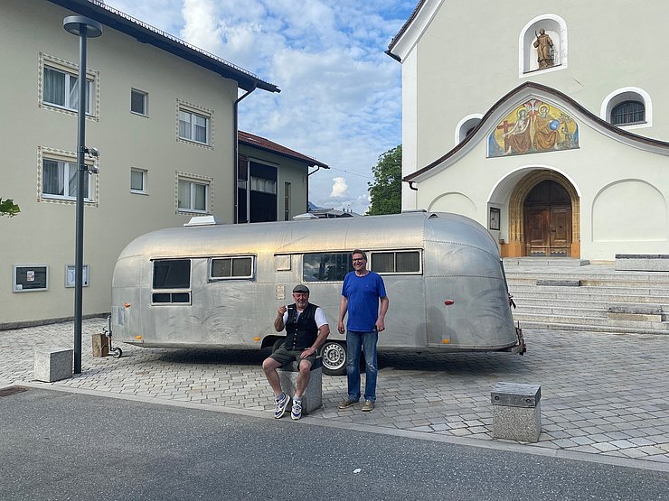 © Alexander Erler Zwei Personen posieren auf einem Platz in Wattens vor dem silbernen Aluminium-Wohnwagen der 1950er-Jahre. Eine Person sitzt auf einem niedrigen Steinblock, die andere steht daneben. Im Hintergrund ist die Kirche alte Pfarrkirche Wattens zu sehen.