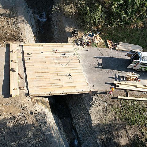 Luftaufnahme der neuen Brücke aus Holz; daneben ein Auto der Baufirma; neben der BRücke liegen noch Werkzeuge und Bretter