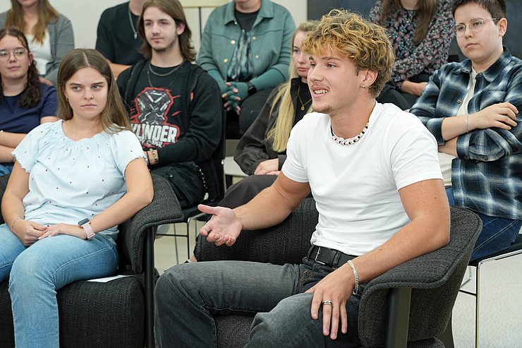 © Land Tirol/ Saxl Ein Schüler sitzt auf einem Stuhl und spricht. Dahinter sitzen weiter Schüler und hören ihm aufmerksam zu.