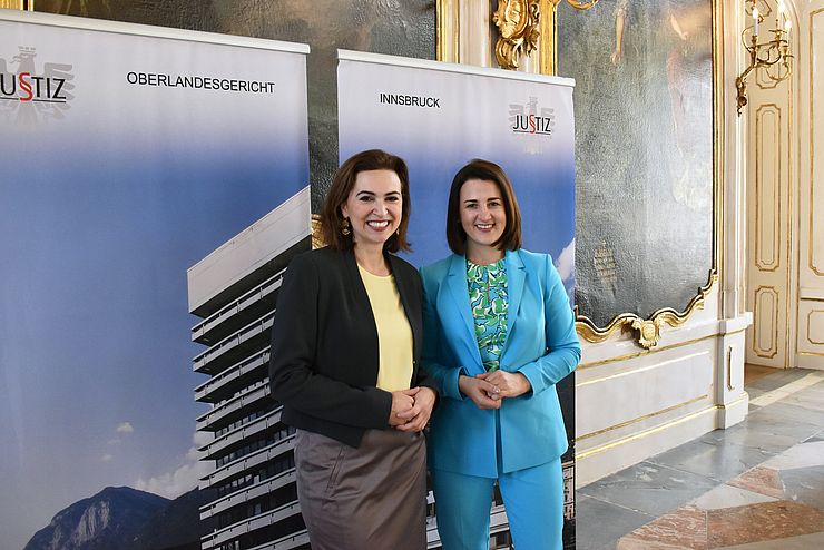 Zadic mit gelber Bluse und schwarzen Blazer und Mair mit Blauen Anzug nebeneinander stehend und in die Kamera lächelnd; im Hintergrund ein Roll-Up mit einem Bild des Oberlandesgericht Innsbruck und der Aufschrift Justiz