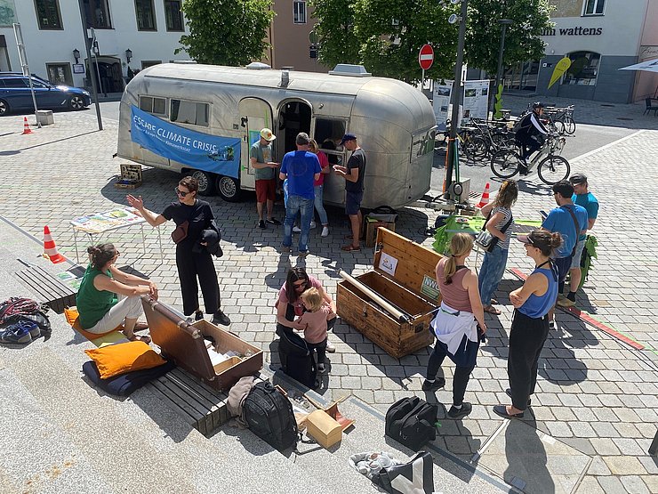 © Alexander Erler Mehrere Menschen treffen sich bei sonnigem Wetter auf einem Platz in Wattens rund um den Aluminium-Wohnwagen der 1950er-Jahre. Am Wagen hängt ein blaues Banner mit der Aufschrift „Escape Climate Crisis“. Auf dem Boden liegen Holzkisten, Taschen, Sitzkissen und Informationsmaterial.