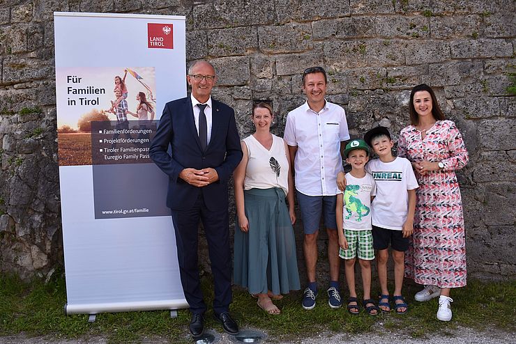 LH Mattle und LRin Mair mit vierköpfiger Familie vor Mauer und Land-Tirol-Rollup