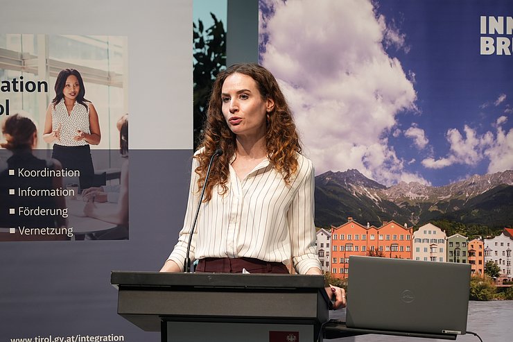 © Land Tirol/Saxl Judith Kohlenberger mit langen braunen Locken und gestreifter Bluse spricht am Rednerpult. Im Hintergrund sind ein Informationsbanner zur Integration in Tirol und ein Banner mit Innsbrucker Altstadt-Panorama zu sehen.