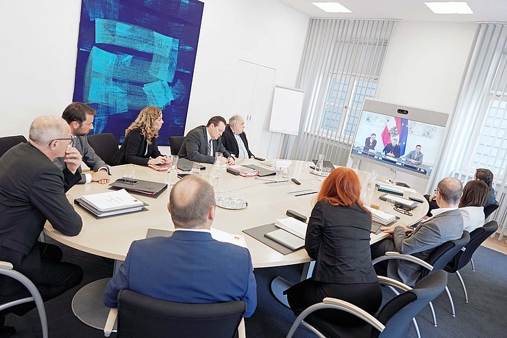 Mehrere Menschen sitzen an einem großen Tisch und blicken auf einen Bildschirm.