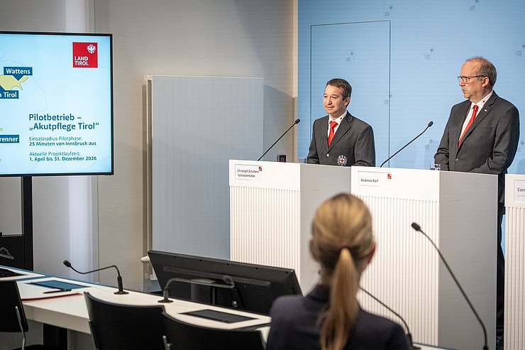 Zwei Männer stehen an einem Rednerpult, links sieht man einen Bildschirm. 