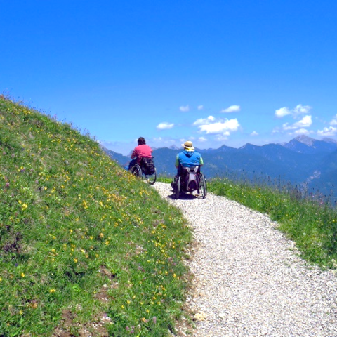 Zwei Rollstuhlfahrer bewegen sich auf einem geschotterten Wanderweg mit Blick in die Bergwelt.