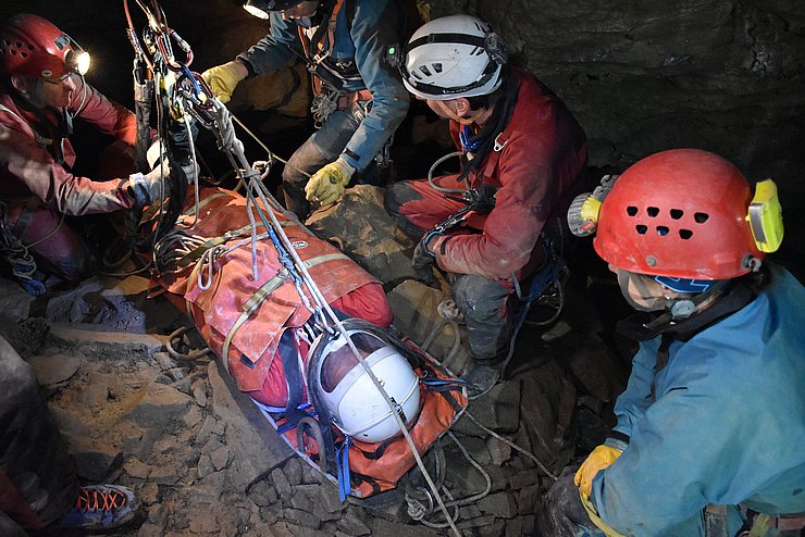 In Höhle: Person in Trage, HelferInnen mit Helmen und Stirnlampen vergurten Trage