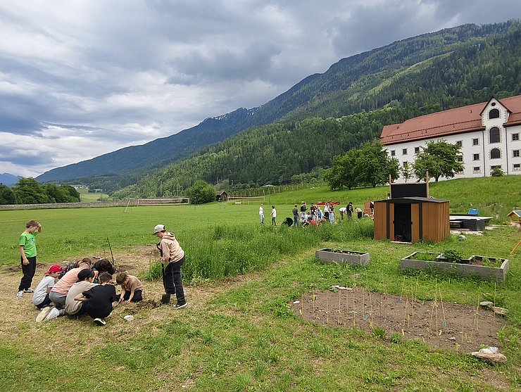 Eine Gruppe von Kindern arbeitet in einem Schulgarten auf einer großen Wiese: Mehrere Kinder knien um ein kleines Erdloch und graben gemeinsam, während andere daneben stehen und zuschauen. Im Hintergrund sind weitere Gruppen bei Gartenarbeiten, Hochbeete, ein kleines Holzhaus sowie ein großes weißes Gebäude zu sehen. Dahinter erheben sich grüne Berghänge unter einem bewölkten Himmel.