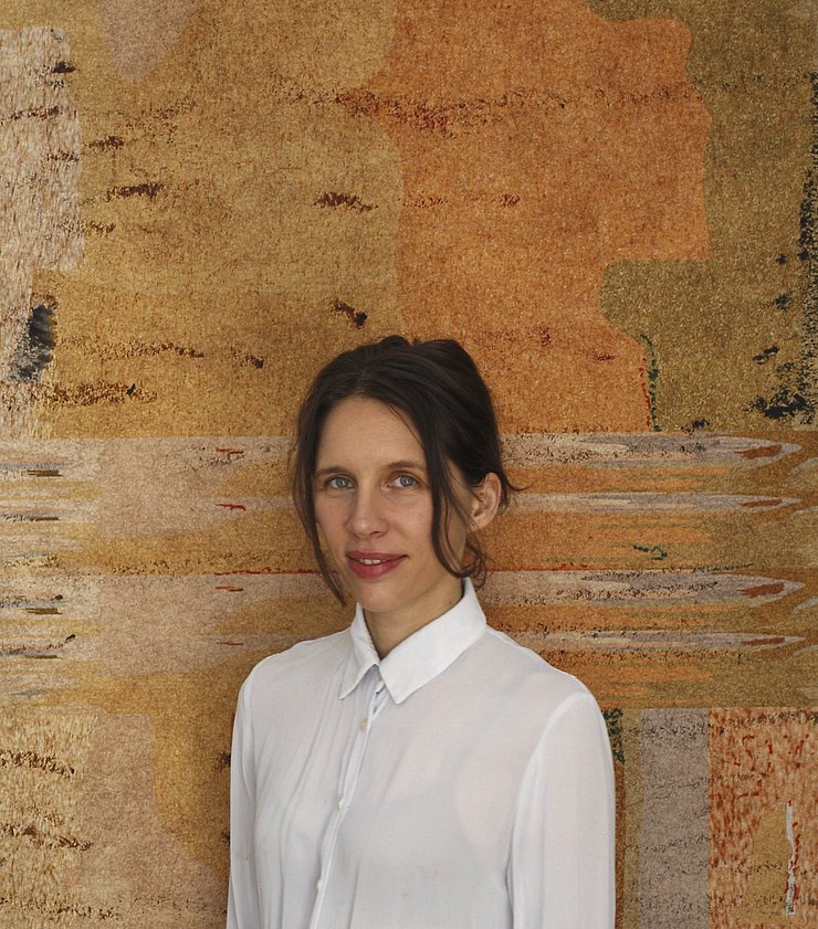 Eine Frau mit zusammengebundenen Haaren steht vor einer bemalten Leinwand. 