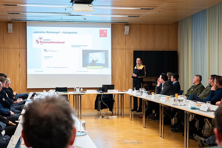 Staudacher vor Publikum stehend und sprechend; Leinwand mit Powerpoint-Präsentation