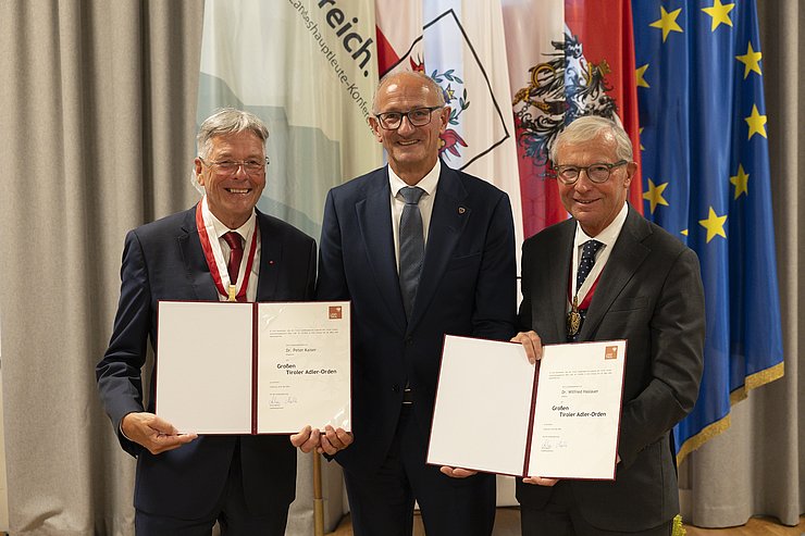 © Land Tirol/Sedlak Drei Männer stehen vor Fahnen, zwei halten Urkunden in die Höhe.