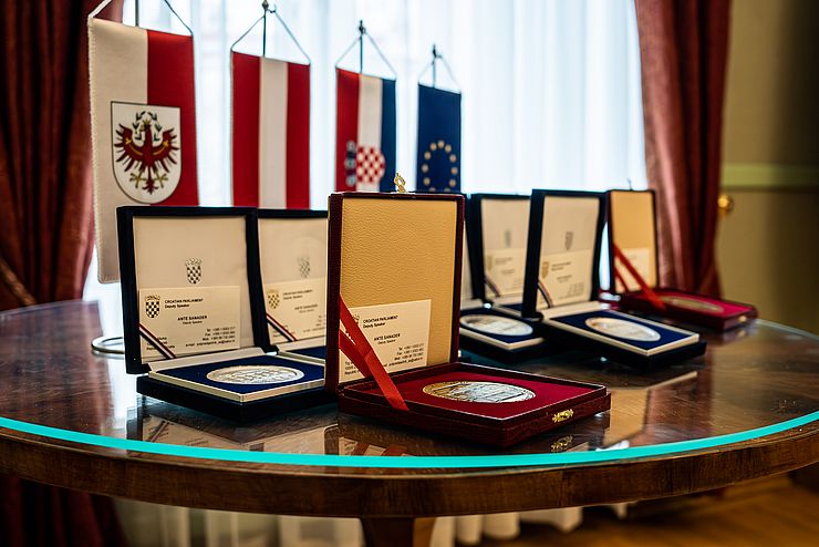 Die Auszeichnungen in roten und blauen Schächtelchen auf einem Tisch liegend. Dahinter vier kleine Fahnen von Tirol. Österreich, Kroatien und der EU