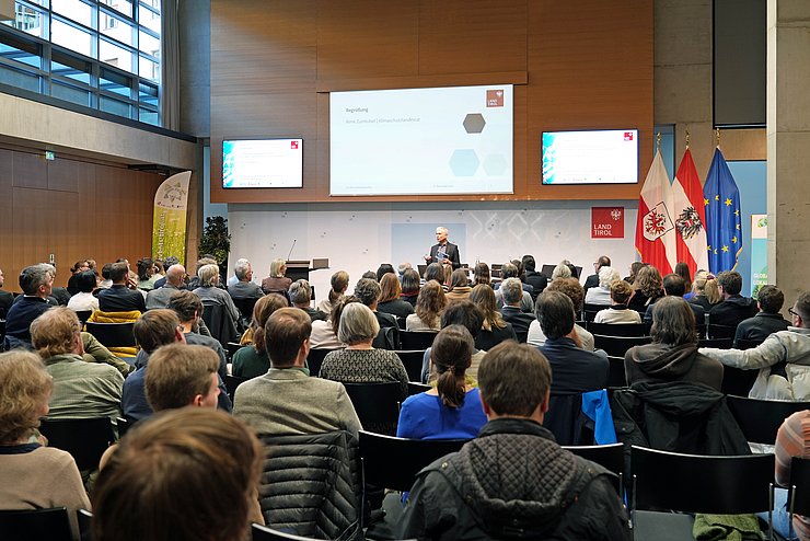 Zahlreiche Menschen sitzen in einem großen Saal und blicken Richtung Bühne.
