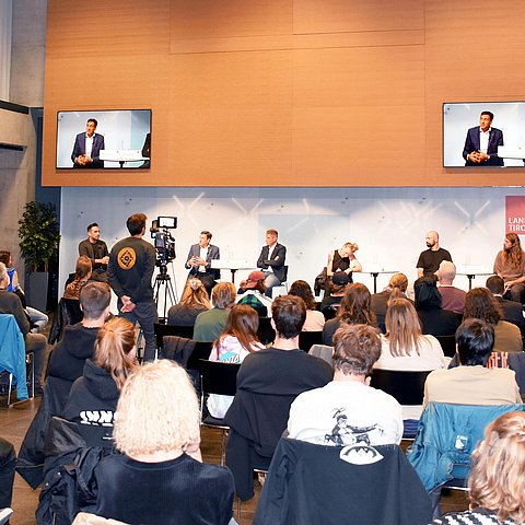 Die TeilnehmerInnen sitzen auf Stühlen im Saal, vorne das Podium