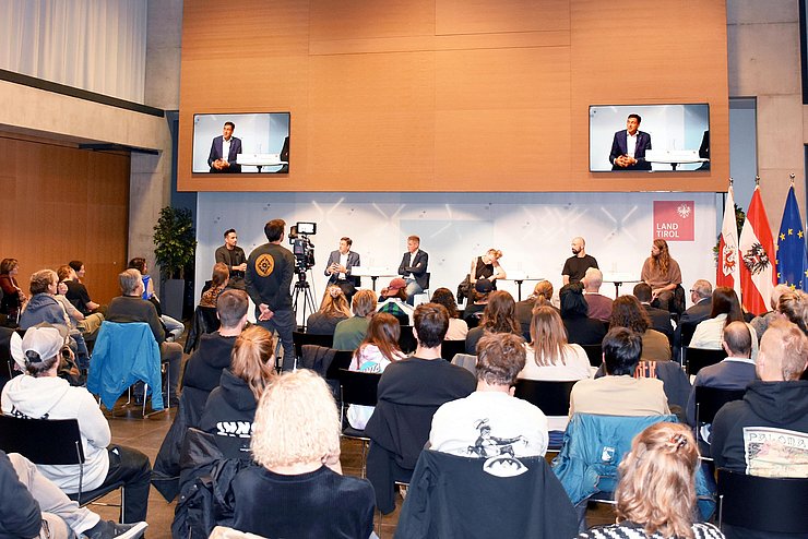 Die TeilnehmerInnen sitzen auf Stühlen im Saal, vorne das Podium