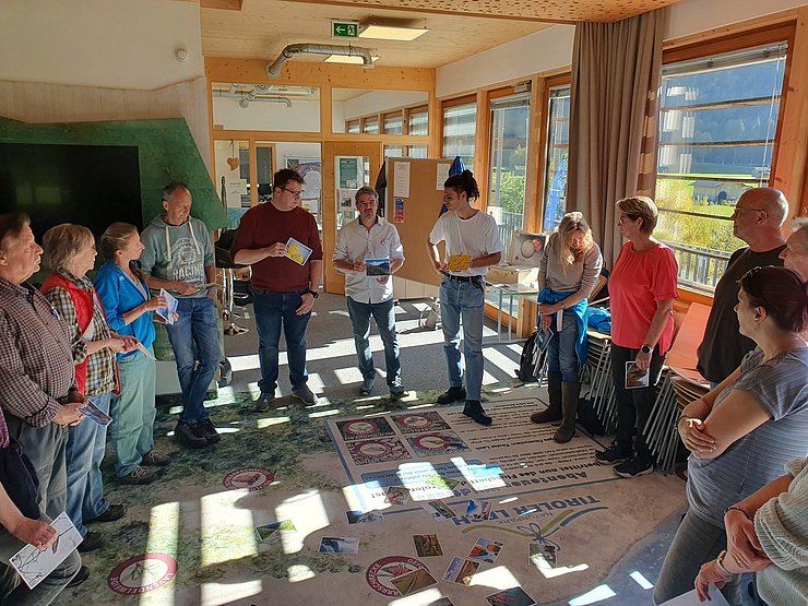 Eine Gruppe von Menschen steht in einem Halbkreis. Sie halten Karten in den Händen. Auf dem Boden liegt eine große bedruckte Plane mit Graphiken und Texten, darauf liegen Bildkarten verteilt.