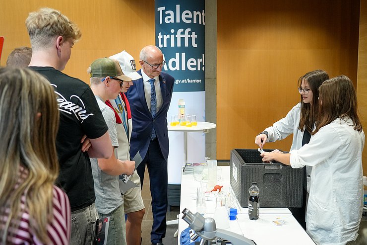 © Land Tirol/Saxl Schülerinnen und Schüler stehen gemeinsam mit Landeshauptmann Anton Mattle vor einem Messestand der Labortechnik und schauen sich das Experiment an.