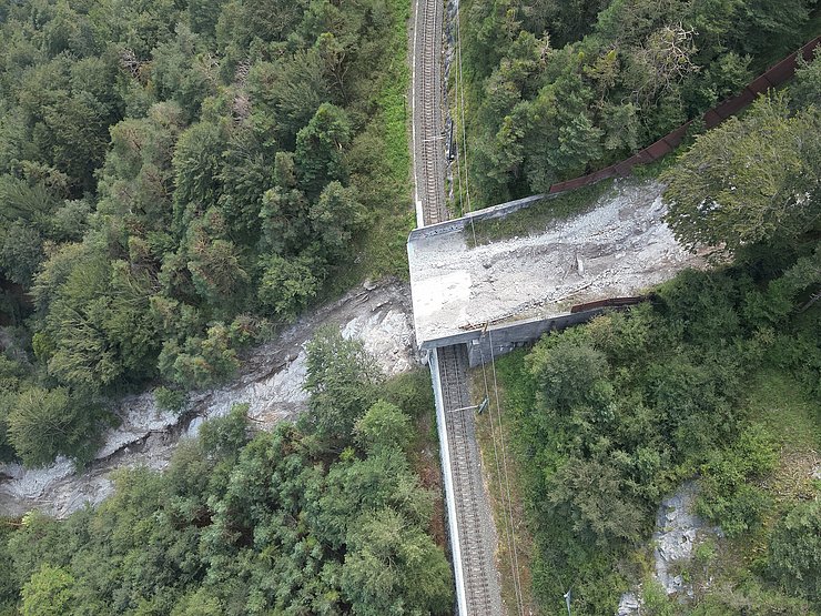 © Land Tirol/Drohnen-Einsatz-Gruppe Blick von oben auf Bahnstrecke in Wald mit Galerie. Auf der Galerie sowie im Wald unterhalb der Galerie sind Ablagerungen eines Murganges zu sehen.