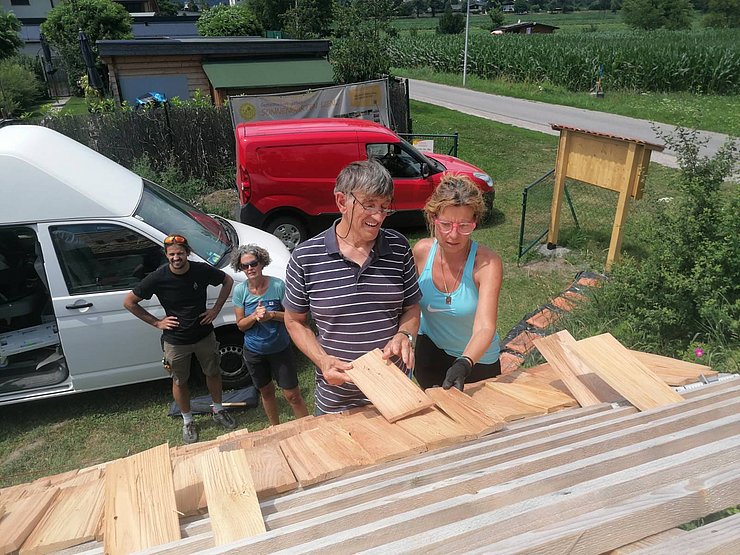 Mehrere Personen arbeiten gemeinsam an dem Dach aus Holzschindeln. Zwei Personen stehen auf dem Dach und legen die rechteckigen Holzschindeln nebeneinander. Weitere Personen stehen darunter und schauen zu.