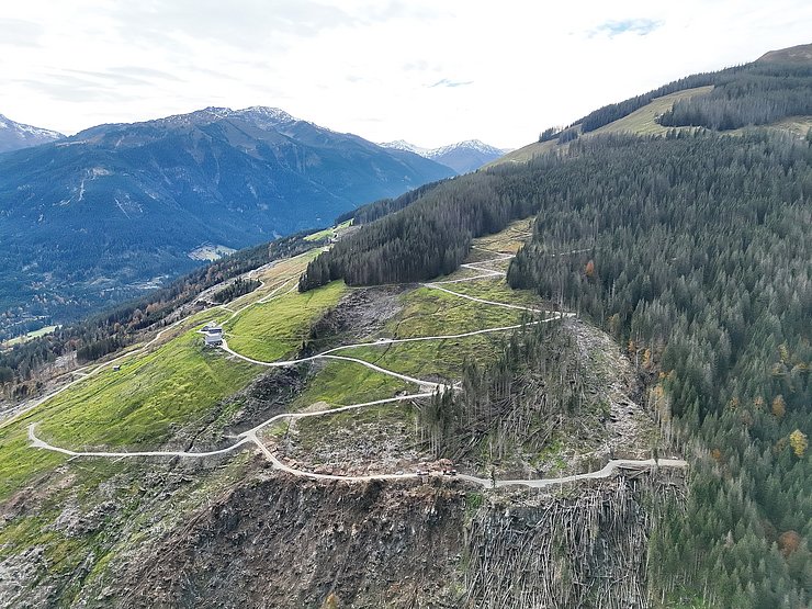 © Land Tirol/Drohnen-Einsatz-Gruppe Blick auf Hang bestehend aus Wiesen- und Waldflächen, durchzogen von einer Fahrstraße. Ein Teil der Waldfläche besteht aus umgestürzten Bäumen, die teilweise bereits weggeräumt wurden.