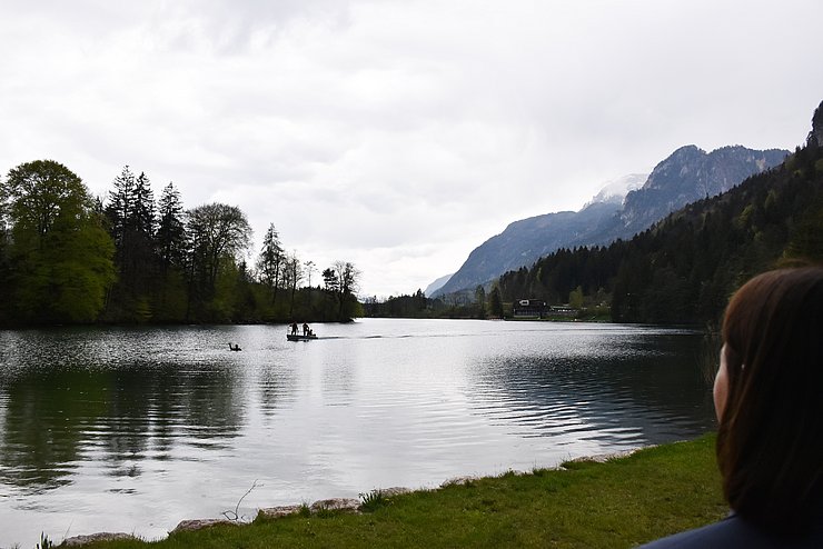 Rechts am Bild der Hinterkopf von Mair; sie blickt auf den Reintalersee vor ihr. Im See schwimmt eine Person und winkt um Hilfe. Ein Boot mit Mitgliedern der Wasserrettung nähert sich. Im Hintergrund der wolkenverhangene Himmel und Berge