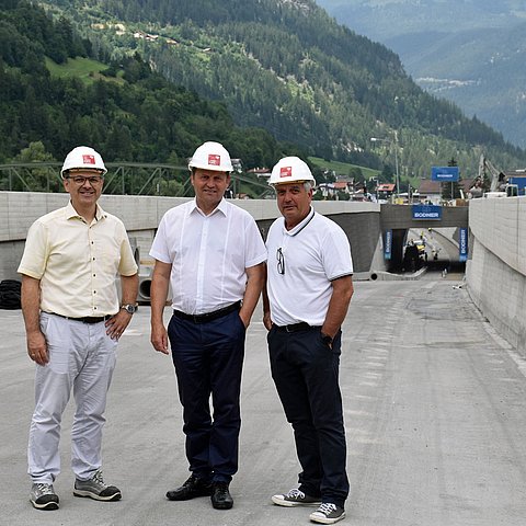 Sommerlich gekleidet stehen vor der Baustelle einer Unterflurtrasse, die bereits deutlich erkennbar ist, von links Baubezirksamtsleiter Bernd Stigger, LHStv Josef Geisler und Bgm Heinz Kofler.